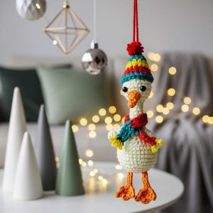May include: A handmade crochet goose Christmas ornament. The goose wears a colorful hat, a red and teal scarf, and has orange feet. The ornament is suspended by a red string, with Christmas decorations in the background.