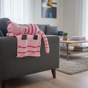 May include: A pink and red crocheted cat-shaped pillow and remote control holder rests on a gray sofa. The cat has striped details and is lying on a pink blanket with pockets for remotes. The room has a modern design.