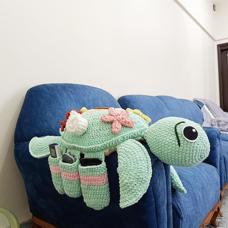 May include: A crocheted turtle-shaped organizer in shades of green, pink, and white, with pockets for storage. The turtle is decorated with flower appliqu&eacute;s and is placed on a blue sofa.