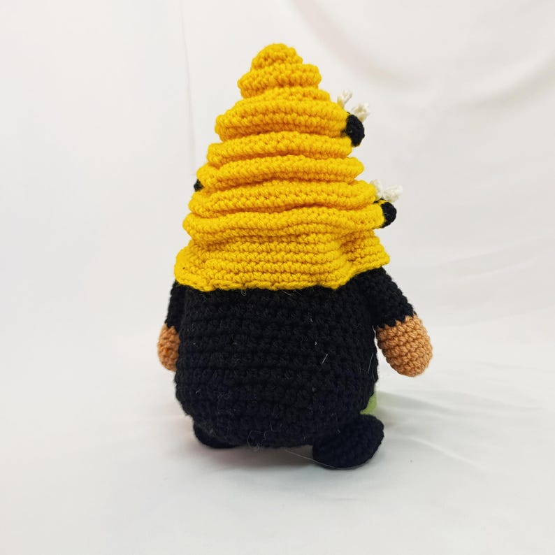 May include: A handmade crochet gnome with a yellow beehive hat and black body. The gnome has small brown hands and feet, and two small bees are attached to the hat. The gnome is a whimsical decorative item.
