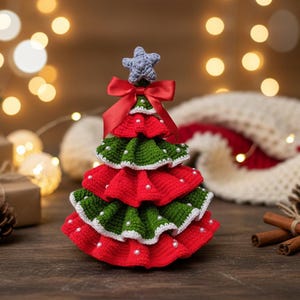 May include: A crocheted Christmas tree ornament. The tree is made with red and green yarn, decorated with white bead details, and topped with a blue star. A red ribbon bow sits beneath the star. The tree is on a wooden surface.