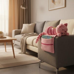 May include: A pink crocheted cat-shaped armrest organizer with a pocket, resting on a gray sofa. The cat has striped details and a smiling face. The scene includes a wooden side table and a window.