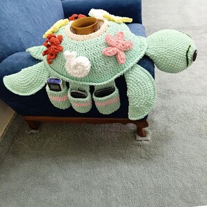 May include: A crocheted turtle-shaped sofa arm organizer in shades of green, with a brown cup holder and pockets for remote controls. The turtle has decorative coral, starfish, and a snail. The organizer is placed on a blue sofa.