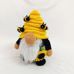 May include: A handmade crochet gnome with a yellow beehive hat and three small bees. The gnome has a white beard, a tan nose, and a sunflower on its body. The gnome's arms and legs are black and tan.