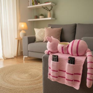 May include: A pink crocheted cat-shaped couch caddy with striped details, holding two remote controls. The caddy has a cup holder and is draped over the arm of a gray sofa. A round jute rug is on the floor.
