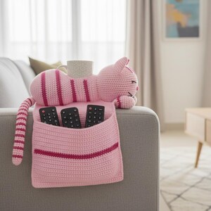 May include: A pink crocheted cat-shaped couch organizer with a pocket for remote controls. The cat has dark pink stripes and a long tail. A mug sits on the cat's back. The organizer is on a gray couch.