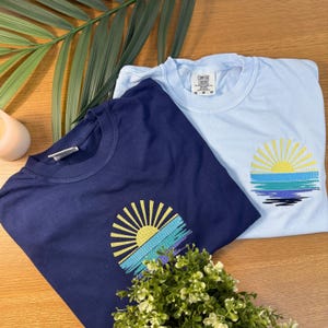 two t-shirts with sun-like embroidered designs, laid out on a wooden surface alongside a green plant and a candle.