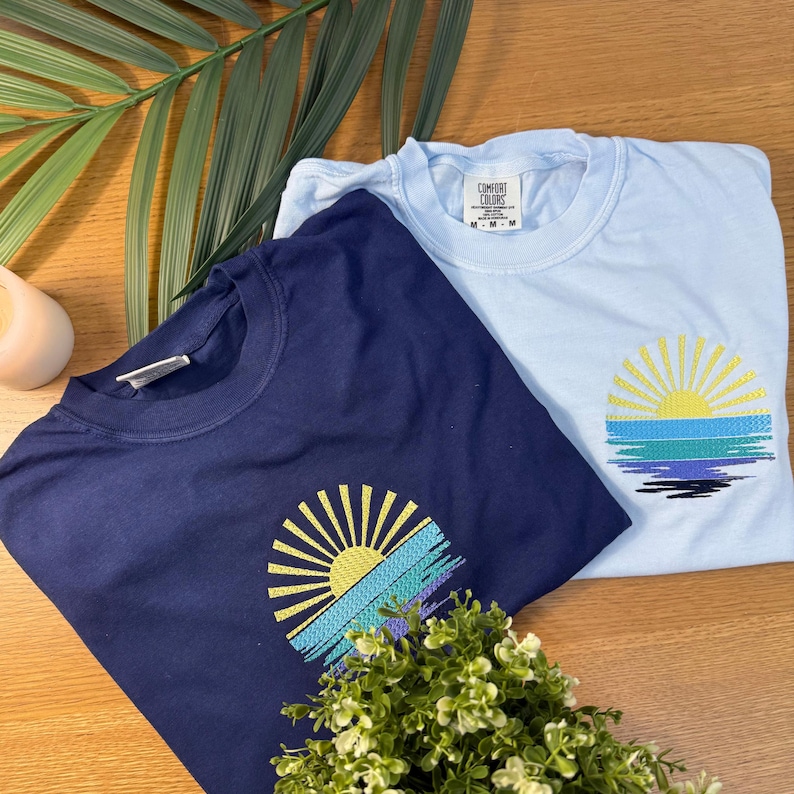 two t-shirts with sun-like embroidered designs, laid out on a wooden surface alongside a green plant and a candle.