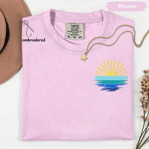 A pink t-shirt with a sun embroidered design, a gold star pendant, and a gold chain necklace are displayed on a white background.