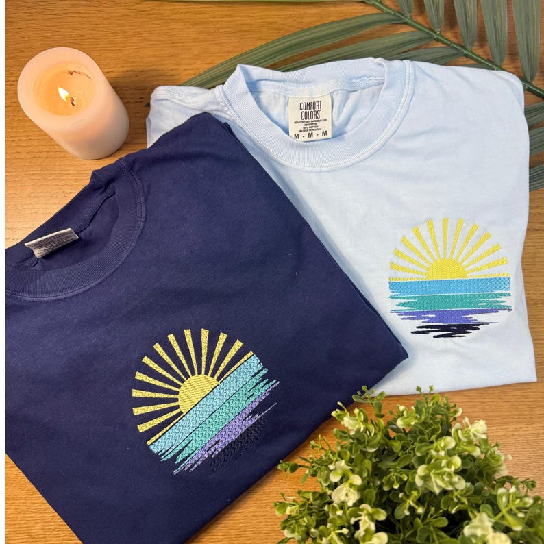 two t-shirts with sun-like embroidered designs, one blue and one white, placed on a wooden surface alongside a lit candle and a potted plant.