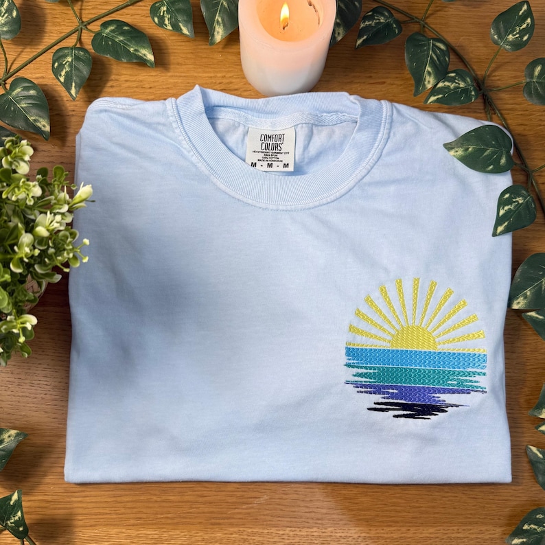 A light blue t-shirt with a sun embroidered design is displayed on a wooden surface, accompanied by a lit candle and green leaves.