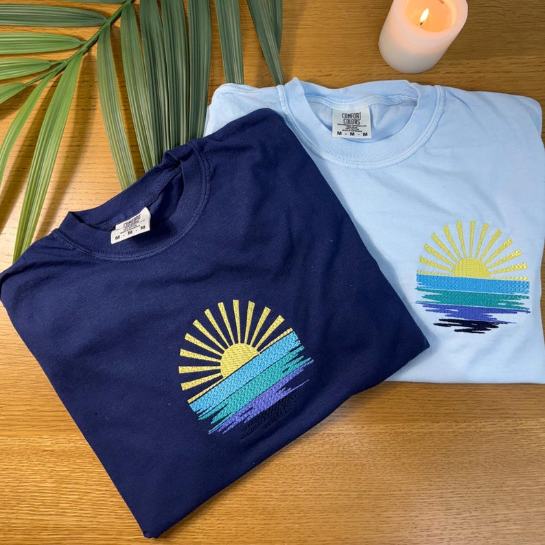 two t-shirts with sun-like embroidered designs, one in navy blue and the other in light blue, placed on a wooden surface alongside a lit candle.
