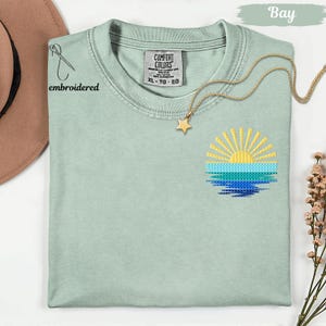 a light green t-shirt with a sun embroidered design on the front, along with a gold star pendant and a brown hat.