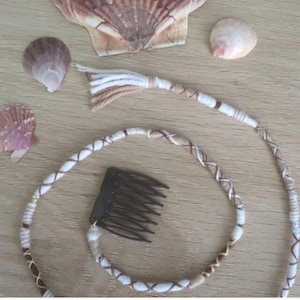 Puede incluir: Una peineta de color marrón oscuro unida a un cordón largo y flexible envuelto en hilo blanco y tostado. El cordón presenta un patrón trenzado y termina en una borla. Tres conchas marinas de diversos tamaños y formas rodean el accesorio.