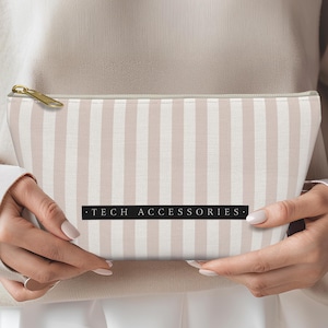 以下が含まれることがあります： ピンクと白のストライプのジッパー付きポーチで、黒い文字で「TECH ACCESSORIES」と印刷されています。
