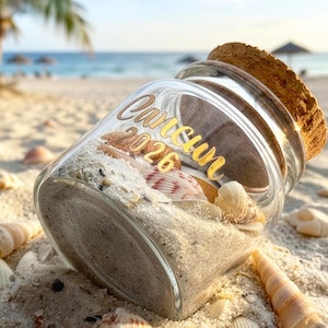Peut inclure: Un bocal en verre transparent avec un bouchon en liège, rempli de sable clair et de divers petits coquillages. L'extérieur du verre présente le texte 'Cancun 2026' imprimé dans une police cursive dorée, situé sur le côté du bocal.
