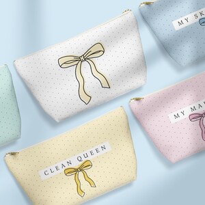 May include: Cosmetic bags in various pastel colors, each adorned with a bow illustration and polka dots. One bag reads "CLEAN QUEEN". Other bags have text labels. The bags have gold zippers.