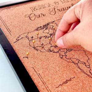 May include: A rectangular cork board featuring a black printed world map and the text "JESSICA & M... Our Travel". The board is framed in black and contains multiple gold and dark-colored push pins inserted into various locations on the map.