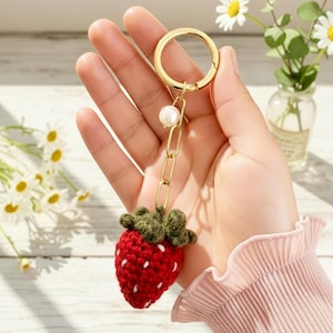 May include: A handmade red crochet strawberry keychain with white seed details and a green leafy top. The keychain has a gold-colored chain, a pearl accent, and a gold-colored clasp.