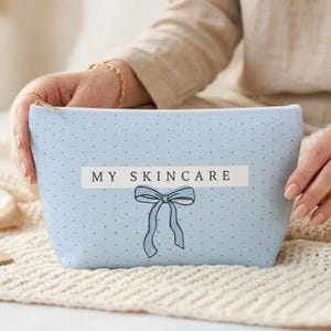 May include: A light blue cosmetic bag with a zipper closure, featuring a polka dot pattern and the text "MY SKINCARE". A bow graphic is below the text. The bag is held by a person.