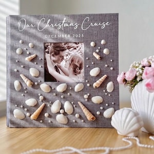 May include: A square photo album with a gray fabric cover featuring the text Our Christmas Cruise and DECEMBER 2025. The cover is decorated with various white seashells and small pearls surrounding a central square photograph of a couple.