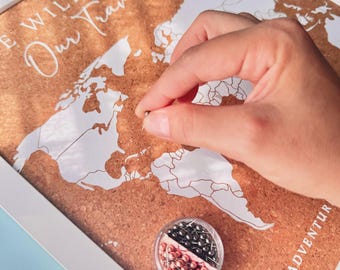 May include: A framed cork board world map with white printed continents and the text "THE WILKINS" and "Our Travel Story" and "LAST ADVENTURE". A small clear container holds silver and rose gold push pins for marking locations on the map.