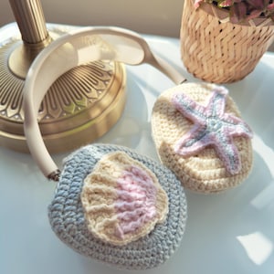 May include: Crocheted headphone covers in light blue and cream, adorned with a seashell and starfish design. The seashell is pink and cream, and the starfish is pink and blue. The headphones are white and beige.