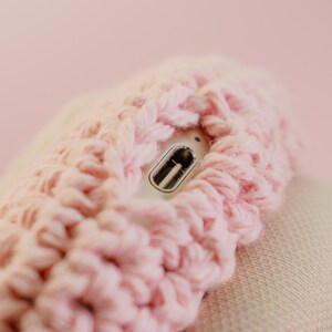 May include: Close-up of a light pink, chunky-knit, crocheted item. A silver and black object is partially visible within the crochet. The background is a soft pink color.