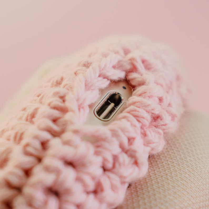 May include: Close-up of a light pink, chunky-knit, crocheted item. A silver and black object is partially visible within the crochet. The background is a soft pink color.