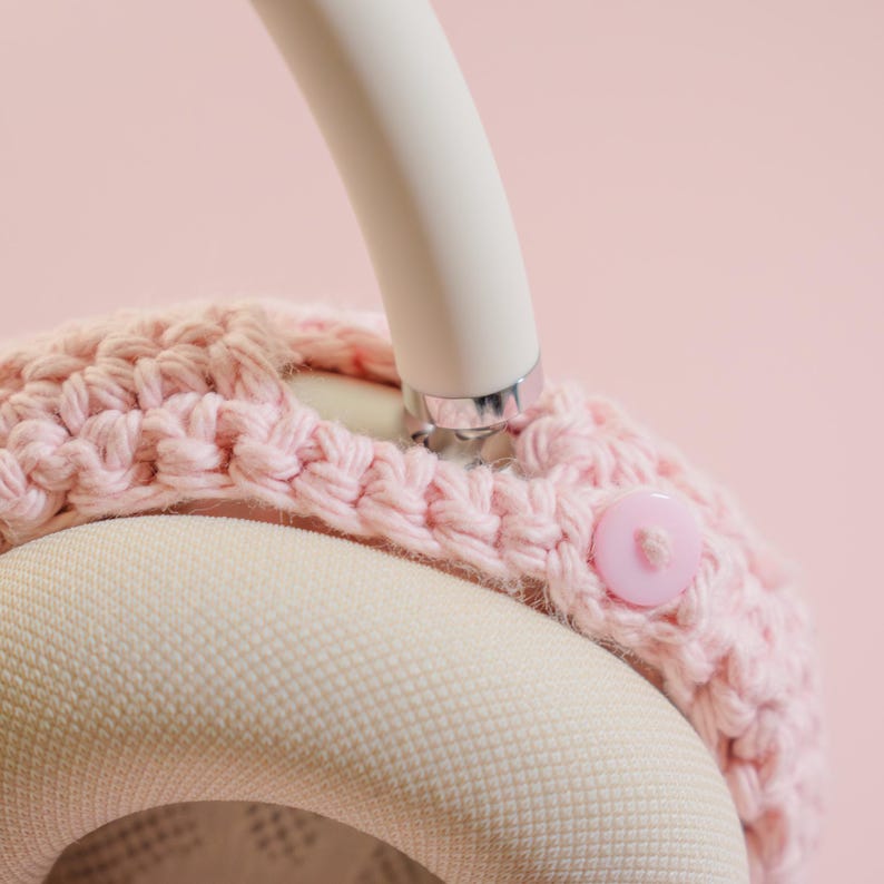 May include: Close-up of a pink crocheted headphone cover with a matching button closure. The cover is placed on a white and silver headphone. The background is a soft pink.