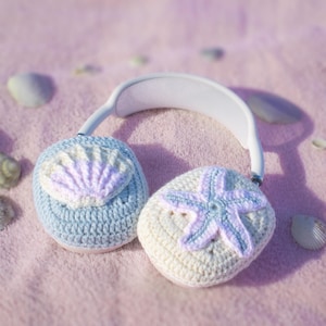 May include: White headphones with crocheted ear covers. One cover is light blue with a seashell design, the other is cream-colored with a starfish. The designs feature pastel colors, evoking a beach theme.