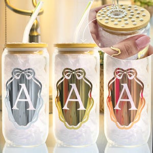 Pode incluir: Três copos de vidro para beber com tampas de bambu e canudos transparentes. Cada copo apresenta um emblema de cor metálica diferente com a letra "A". Os emblemas são prateados, dourados e acobreados. Os copos são preenchidos com papel de seda branco.