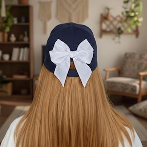 May include: A navy blue baseball cap with a large white bow on the back. The cap is made of a solid, dark blue fabric, and the bow is a contrasting bright white. The image shows the back of the cap, with the bow as the focal point.