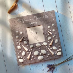 Könnte beinhalten: Ein quadratischer, grauer Bilderrahmen mit Stoffstruktur, verziert mit verschiedenen weißen Muscheln und kleinen Perlen. Der Rahmen trägt oben den Text 'The Summer I Turned 16' und '2026', mit einem mittigen Einsatz 'Thank you' und 'LUMEVA'.