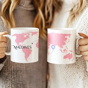 以下が含まれることがあります： ピンクの世界地図のデザインが施された2つの白いセラミックマグ。1つのマグには、点線と飛行機のグラフィックが付いた「モルディブ」の文字が描かれています。もう1つのマグには、青いハートのグラフィックと点線があります。どちらのマグにもハンドルが付いています。