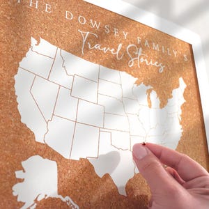 May include: A corkboard map of the United States with white state outlines and the text "THE DOWSEY FAMILY'S Travel Stories." A hand places a red pin on the map. The map is framed in white.