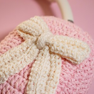May include: Close-up of a handmade, crocheted item in soft pink and cream colors. The item features a large bow design, with the bow and its accents in cream, and the base in pink. The crochet stitches are clearly visible, adding texture.