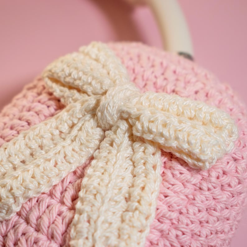 May include: Close-up of a handmade, crocheted item in soft pink and cream colors. The item features a large bow design, with the bow and its accents in cream, and the base in pink. The crochet stitches are clearly visible, adding texture.