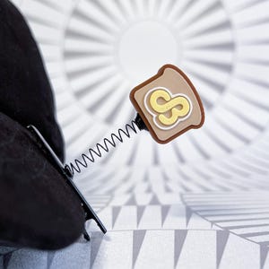 May include: A novelty hair clip featuring a slice of toast with a peanut butter and jelly design. The toast has a brown crust, a tan center, and a yellow "S" design. The clip is attached to a black spring and a black metal clip.