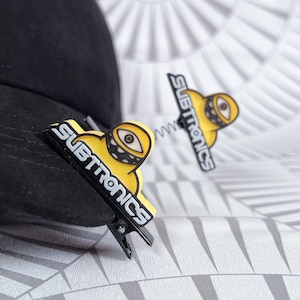 May include: Two yellow and black hair clips with the word "SUBTRONICS" and a cartoon eye design. One clip is attached to a black hat. The clips have a spring-like design connecting the eye to the clip.
