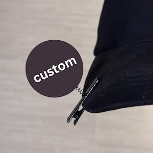 May include: Black hat with a metal clip attached. A dark purple circle with the word "custom" in white text is in the image. The hat appears to be made of a soft material.