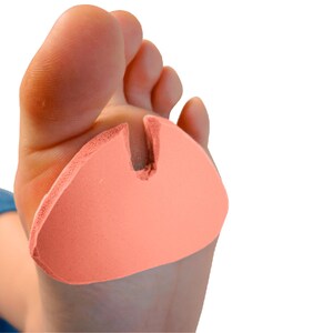 May include: A peach-colored foam metatarsal pad is placed on the foot. The pad has a curved shape with a split to accommodate the toes. The foot is in the foreground, with a white background.