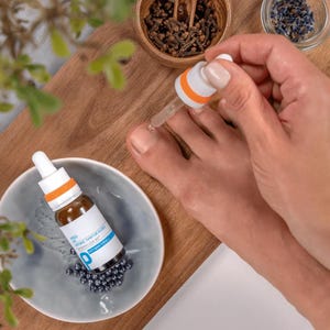 Puede incluir: Una pequeña botella de vidrio marrón con un cuentagotas blanco y naranja se coloca en un plato gris con pequeñas cuentas azul oscuro. El cuentagotas se usa en un dedo del pie. La botella tiene texto blanco y azul.