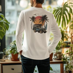 May include: White long-sleeve shirt featuring a tropical graphic on the back. The design includes a vintage blue jeep with surfboards, palm trees, a sunset, and birds. The shirt is made of a soft, comfortable material.
