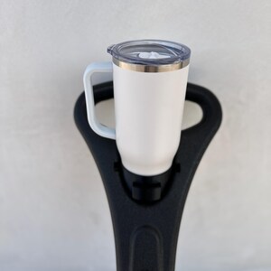 May include: White stainless steel travel mug with a black handle and a clear lid. The mug is sitting on a black holder.
