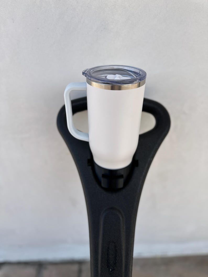 May include: White stainless steel travel mug with a black handle and a clear lid. The mug is sitting on a black holder.