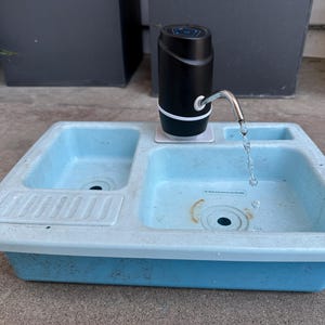 May include: A light blue utility sink with a black water dispenser. The sink has two basins and a small soap dish. Water flows from the dispenser's spout into the sink. The dispenser is black with a white trim.