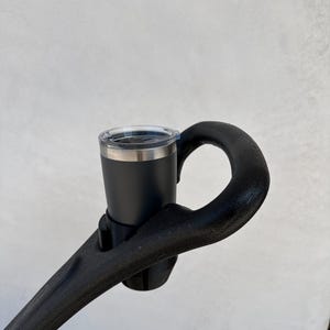 May include: A black cup holder attached to a black handlebar. The cup holder is holding a black travel mug with a clear lid.