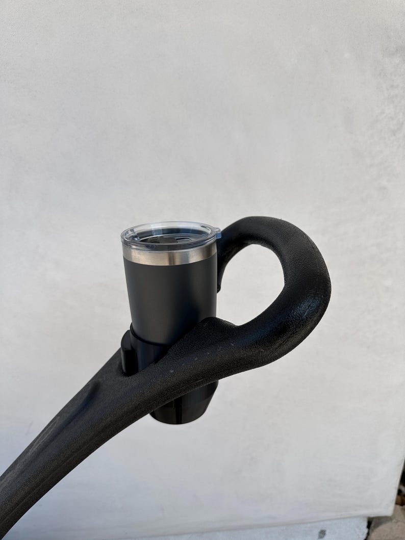May include: A black cup holder attached to a black handlebar. The cup holder is holding a black travel mug with a clear lid.