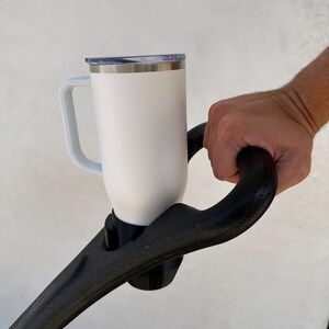 May include: White travel mug with a black handle, attached to a black stroller handle.
